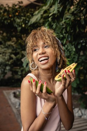 Positive junge afrikanisch-amerikanische Frau mit Hosenträgern im Sommerkleid, die frische Papaya in der Hand hält und wegschaut, während sie neben verschwommenen Pflanzen in der Orangerie steht, inspiriert von tropischen Pflanzen, Sommerkonzept
