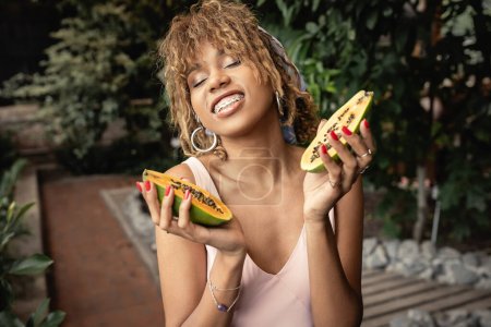 Smiling young african american woman with braces waring summer outfit and closing eyes while holding fresh papaya in blurred indoor garden, fashion-forward lady inspired by tropical plants
