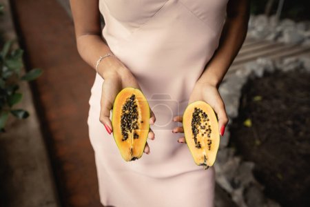 Ausgeschnittene Ansicht der trendigen jungen afrikanisch-amerikanischen Frau im Sommerkleid mit frisch geschnittener Papaya im Stehen im Gartencenter, modebewusste Dame inspiriert von tropischen Pflanzen, Sommerkonzept