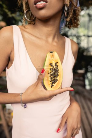 Vista recortada de la elegante joven afroamericana con tirantes que usan vestido de verano mientras sostiene papaya madura cortada en el centro del jardín borroso, dama de moda hacia adelante inspirada en las plantas tropicales