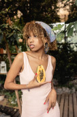 Fashionable and young african american woman in headscarf and summer dress holding cut ripe papaya while standing near plants in greenhouse, fashion-forward lady inspired by tropical plants puzzle #663911840