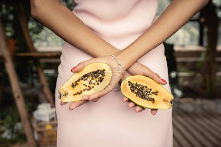 Vista recortada de la joven afroamericana en vestido de verano sosteniendo papaya fresca y de pie en el jardín interior borroso, mujer de moda rodeada de exuberancia tropical