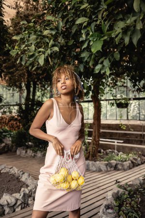 Mujer afroamericana joven de moda en pañuelo para la cabeza y vestido de verano sosteniendo bolsa de malla con limones frescos mientras está de pie en el centro del jardín, mujer de moda rodeada de exuberancia tropical, concepto de verano