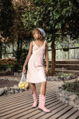 Trendy young african american woman in summer dress holding fresh lemons in mesh bag and standing in blurred indoor garden at background, stylish lady enjoying tropical atmosphere magic mug #663911908