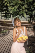 Pleased young african american woman in summer dress holding mesh bag with fresh lemons and looking at camera near plants in orangery, stylish lady enjoying tropical atmosphere, summer concept mug #663911922