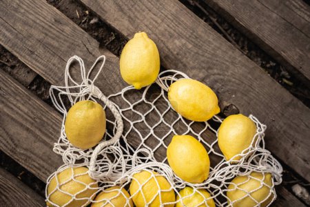 Top view of fresh and juicy lemons lying near mesh bag on wooden planks, yellow fruit, citrus, composition, ingredients, rustic, sour food, vitamin c, summer concept 
