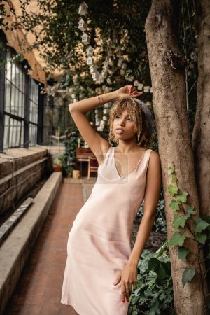 Jeune femme afro-américaine branchée en foulard et robe d'été relaxante tout en se tenant près des arbres dans un jardin intérieur flou, femme élégante avec des plantes tropicales en toile de fond, concept d'été