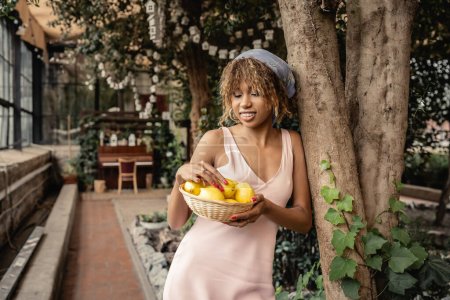 Alegre mujer afroamericana con tirantes en pañuelo para la cabeza y vestido de verano mirando limones frescos en la cesta y de pie cerca de los árboles en el jardín interior, mujer elegante con plantas tropicales en el telón de fondo