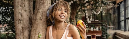 Mujer afroamericana joven y de moda con tirantes y ropa de verano sosteniendo limón maduro y de pie cerca de los árboles en el centro del jardín, señora de la moda hacia adelante en armonía con la flora tropical, pancarta 