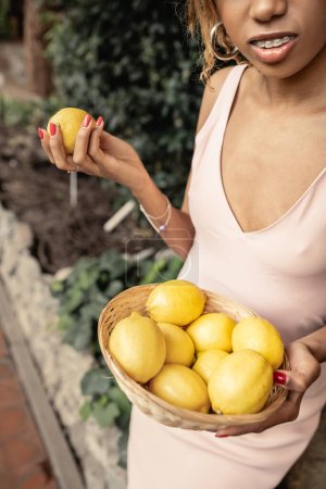 Ausgeschnittene Ansicht einer jungen afrikanisch-amerikanischen Frau mit Hosenträgern im Sommerkleid und mit saftigen Zitronen im Korb im Gartencenter, modebewusste Dame im Einklang mit tropischer Flora