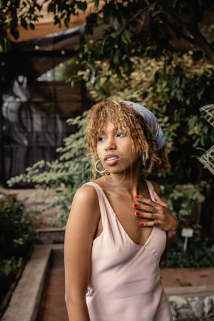 Portrait of stylish young african american woman in headscarf and summer dress touching chest and standing in blurred indoor garden, woman in summer outfit posing near lush tropical plants