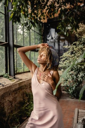 Foto de Relajada joven afroamericana mujer en traje de verano y pañuelo en la cabeza tocando el cuello mientras está de pie cerca de las plantas en el jardín interior, mujer en traje de verano posando cerca de exuberantes plantas tropicales - Imagen libre de derechos