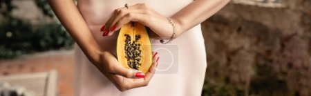 Photo for Cropped view of african american woman in stylish summer dress holding cut and juicy papaya while standing in garden center, woman in summer outfit posing near lush tropical plants, banner - Royalty Free Image