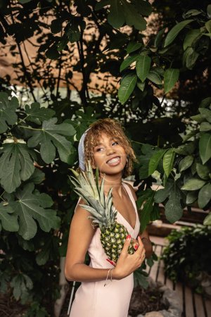 Smiling young african american woman with braces wearing summer outfit and holding pineapple and standing near plants in orangery, woman in summer outfit posing near lush tropical plants