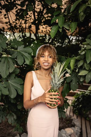 Young and cheerful african american woman with braces wearing summer outfit and holding pineapple while standing near plants in orangery, woman in summer outfit posing near lush tropical plants