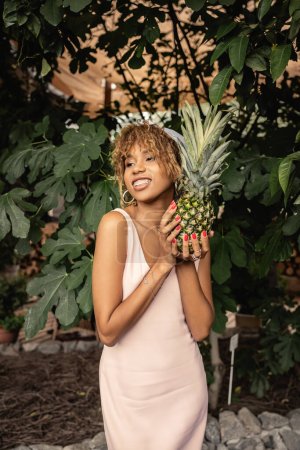 Agréable femme afro-américaine avec bretelles en tenue d'été et tenant l'ananas debout dans le jardin intérieur en arrière-plan, femme en tenue d'été posant près de plantes tropicales luxuriantes