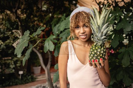 Téléchargez les photos : Joyeux jeune femme afro-américaine avec bretelles en tenue d'été tenant l'ananas juteux et regardant la caméra en orangerie floue, femme en tenue d'été posant près de plantes tropicales luxuriantes - en image libre de droit