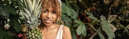 Photo for Positive young african american woman with braces and summer outfit looking at camera near fresh pineapple in blurred garden center, woman in summer outfit posing near lush tropical plants, banner - Royalty Free Image