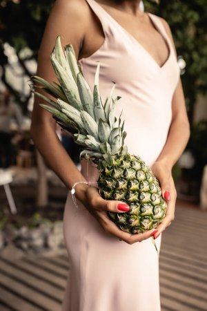 Vue recadrée d'une jeune femme afro-américaine floue en robe d'été tenant de l'ananas frais et debout dans une orangerie floue, femme élégante portant une tenue d'été entourée de feuillage tropical