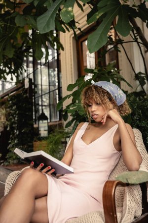 Trendige afrikanisch-amerikanische Frau im Sommer-Outfit liest Buch, während sie auf einem Sessel in der Orangerie sitzt, stilvolle Frau im Sommer-Outfit umgeben von tropischem Laub, Sommerkonzept