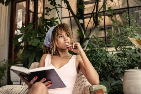 Junge und selbstbewusste Afroamerikanerin im Sommer-Outfit, Buch in der Hand und auf Sessel im verschwommenen Indoor-Garten sitzend, stilvolle Frau im Sommer-Outfit umgeben von tropischem Laub