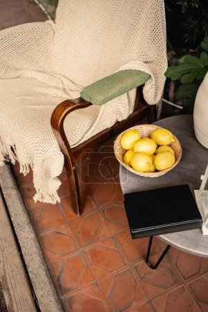 Vista de alto ángulo del libro y limones frescos en un tazón cerca de un sillón y plantas borrosas en naranjería, vitamina c, comida agria, frutas dentro del jardín interior, concepto de verano