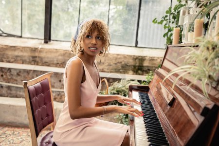 Smiling young african amrican woman in summer dress and headscarf playing piano while spending time in modern indoor garden, woman in comfortable and trendy summer outfit