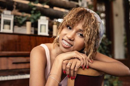 Portrait of smiling and trendy african american woman with braces looking at camera while sitting near blurred piano in indoor garden, woman in comfortable and trendy summer outfit