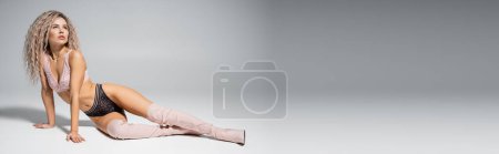 volle Länge der ausdrucksstarken Frau in Spitzenunterwäsche und hohen Stiefeln auf grauem Hintergrund sitzend und wegschauend, sexy und schlanker Körper, gefärbte aschblonde Haare, Mode und Stil, Banner mit Kopierraum