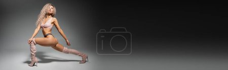 Téléchargez les photos : Longueur totale de femme fashion-forward et sexy en lingerie dentelle et bottes longues debout en pose expressive sur fond noir et gris, corps tonique, cheveux blonds cendrés ondulés, bannière avec espace de copie - en image libre de droit