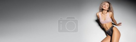 femme sexy avec les yeux fermés et les cheveux blonds frêne ondulé assis dans une pose provocante sur fond gris, sous-vêtements en dentelle, corps tonique, féminité et style, vue grand angle, bannière avec espace de copie