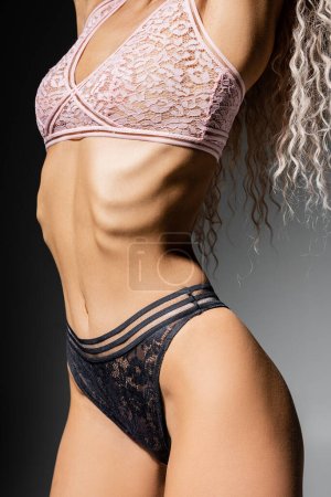 cropped view of woman with toned body and wavy ash blonde hair standing and posing in lace panties and bra on black and grey background, style and femininity, sexy fashion photography