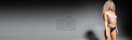 Téléchargez les photos : Séduisante femme au corps tonique et aux cheveux blonds cendrés teints debout sur fond noir et gris et regardant ailleurs, sous-vêtements en dentelle, tendance mode sexy, bannière avec espace de copie - en image libre de droit