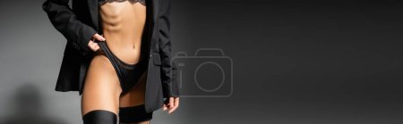 vista recortada de la mujer glamour con el cuerpo tonificado ajustando las bragas mientras posan en chaqueta negro y medias sobre fondo gris, sexualidad y moda, pancarta con espacio para copiar