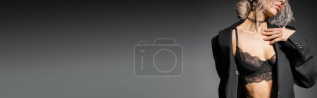 Teilaufnahme einer sinnlichen Frau mit welligem aschblondem Haar, die in SpitzenbH und schwarzem Blazer posiert und die Brust auf grauem Hintergrund berührt, sexy Look, Glamour und Weiblichkeit, Banner mit Kopierraum