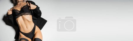 Téléchargez les photos : Confiance et expression de soi, vue partielle de la femme sensuelle avec un corps tonique posé en blazer noir, bas, culotte et soutien-gorge en dentelle sur fond gris, bannière avec espace de copie, vue du dessus - en image libre de droit