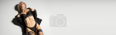 Draufsicht auf attraktive Frau mit durchtrainiertem Körper und welligem aschblonden Haar, in stylischer Dessous, schwarzem Blazer und Strümpfen auf grauem Hintergrund, sexy Modefotografie, Banner mit Kopierplatz