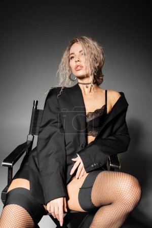 stylisches weibliches Modell in schwarzem Blazer, Dessous und Netzstrümpfen auf Stuhl sitzend und vor grauem Hintergrund wegschauend, welliges aschblondes Haar, ausdrucksstarker Blick, Glamour, sexy Lebensstil