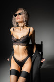 provocative woman with wavy ash blonde hair, in dark sunglasses, sexy lingerie and stockings posing with blazer near chair on black and grey background, individuality and self-expression t-shirt #663914774