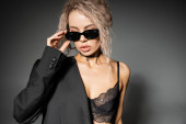 erotic fashion, modern woman with dyed ash blonde hair wearing lace bra and blazer, adjusting dark trendy sunglasses on grey background, glamour style, expressive individuality mug #663914842