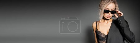 mujer de moda con cabello rubio ceniza teñido, con sujetador de encaje y chaqueta, ajuste de gafas de sol oscuras y de pie sobre fondo gris, aspecto sexy, confianza, pancarta con espacio para copiar