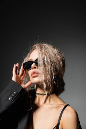 retrato de mujer sexy en chaqueta y collar ajustando gafas de sol oscuras y mirando hacia otro lado sobre fondo negro y gris, pelo rubio ceniza, estilo glamour, confianza, auto-expresión moderna