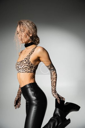 Photo for Seductive woman with slender body and dyed ash blonde hair posing in animal print crop top, long gloves and latex pants while touching black boot on grey background, sexuality and fashion - Royalty Free Image