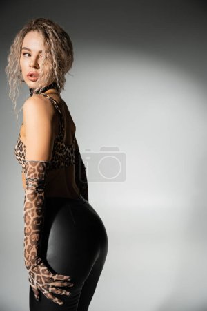 trendy and sexy woman with seductive gaze and wavy ash blonde hair looking away while posing in animal print crop top, long gloves and black latex pants on grey background, sensuality and style