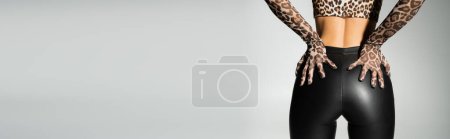 Photo for Sexy fashion trend, partial view of seductive female model in leopard print gloves, crop top and black latex pants touching sexy buttocks while standing on grey background, banner with copy space - Royalty Free Image