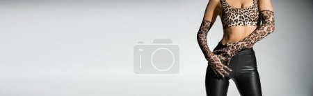 vista recortada de la mujer expresiva y sexy con cuerpo delgado y elegante traje posando en pantalones de látex negro, top de impresión animal y guantes largos sobre fondo gris, pancarta con espacio para copiar