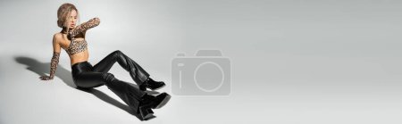 Téléchargez les photos : Longueur totale de femme à la mode et sexy en pantalon en latex noir, haut de culture imprimé léopard et gants longs assis sur fond gris, corps mince, cheveux blonds cendrés ondulés, bannière avec espace de copie - en image libre de droit