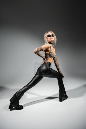 sexy Frau in voller Länge mit dunkler Sonnenbrille, bauchfreiem Top mit Animal-Print, langen Handschuhen und schwarzer Latexhose, die mit der Hand an der Hüfte auf grauem Hintergrund posiert, Glamour-Stil, moderne Selbstdarstellung