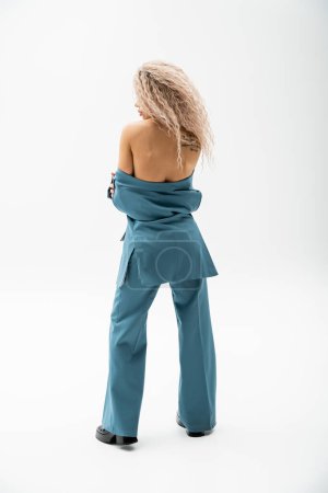 back view of seductive and tattooed woman with bare shoulders wearing blue oversize pantsuit on shirtless body while standing on grey background, glamour fashion, modern self-expression