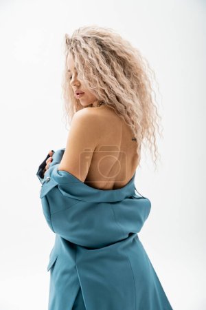 Photo for Erotic and charming woman with wavy ash blonde hair wearing blue blazer on shirtless body while standing on grey background, sexy look, individuality and self-expression - Royalty Free Image